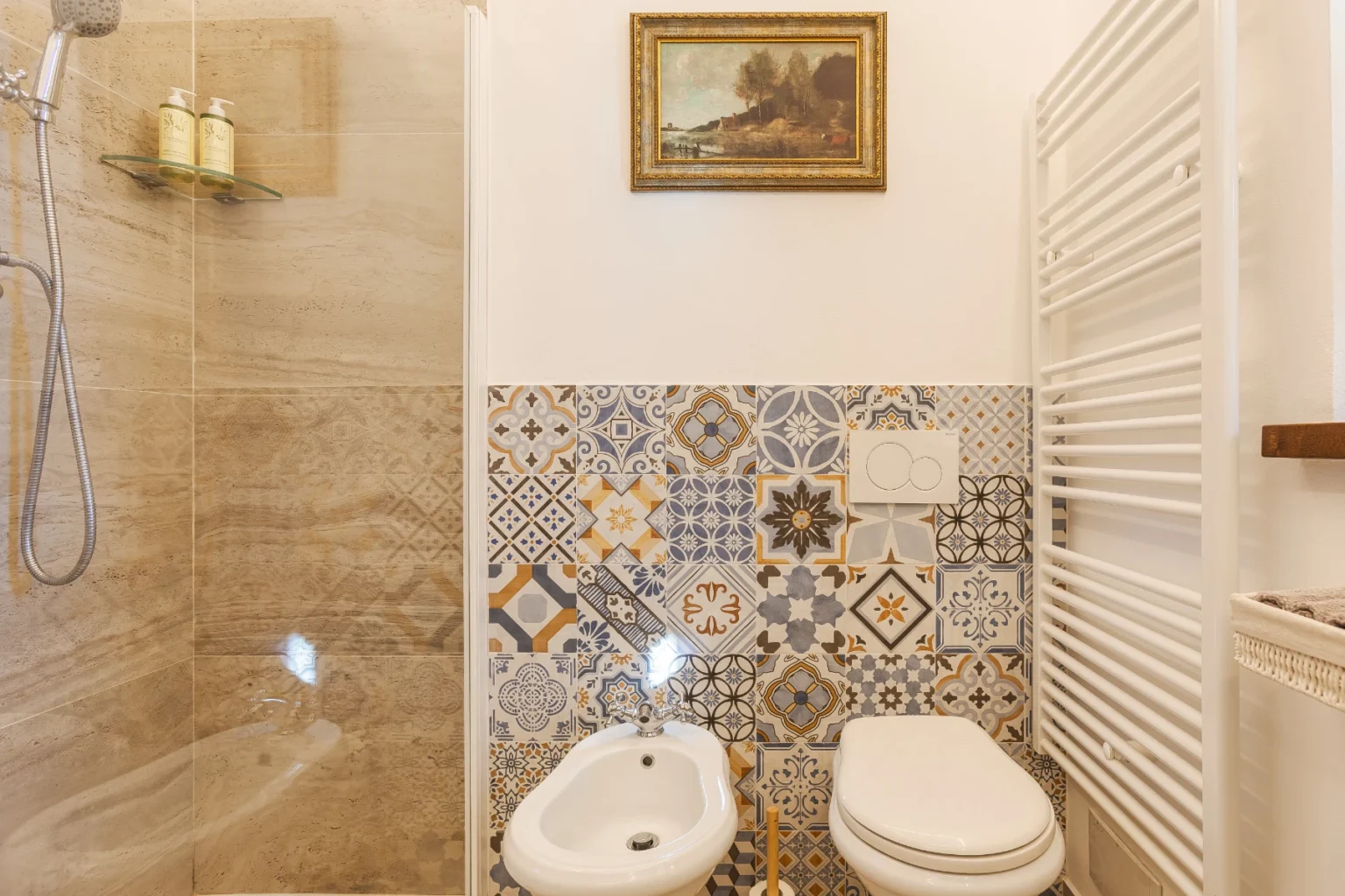 Modern bathroom featuring a walk-in shower with glass enclosure, decorative patterned tiles, a bidet, and a white toilet. A stylish towel radiator and framed artwork enhance the aesthetic appeal. | Only on SopranoVillas