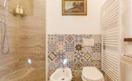 Modern bathroom featuring a walk-in shower with glass enclosure, decorative patterned tiles, a bidet, and a white toilet. A stylish towel radiator and framed artwork enhance the aesthetic appeal.
