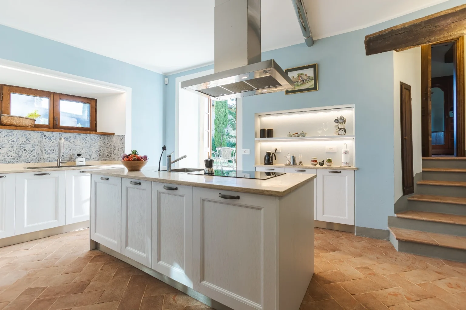 Bright and modern kitchen featuring a central island with a marble countertop, stainless steel range hood, and wooden cabinets. The light blue walls and patterned backsplash create a fresh atmosphere, complemented by natural light from the windows. Decorative items are displayed on open shelves, adding style to the functional space. | Only on SopranoVillas