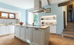 Bright and modern kitchen featuring a central island with a marble countertop, stainless steel range hood, and wooden cabinets. The light blue walls and patterned backsplash create a fresh atmosphere, complemented by natural light from the windows. Decorative items are displayed on open shelves, adding style to the functional space.