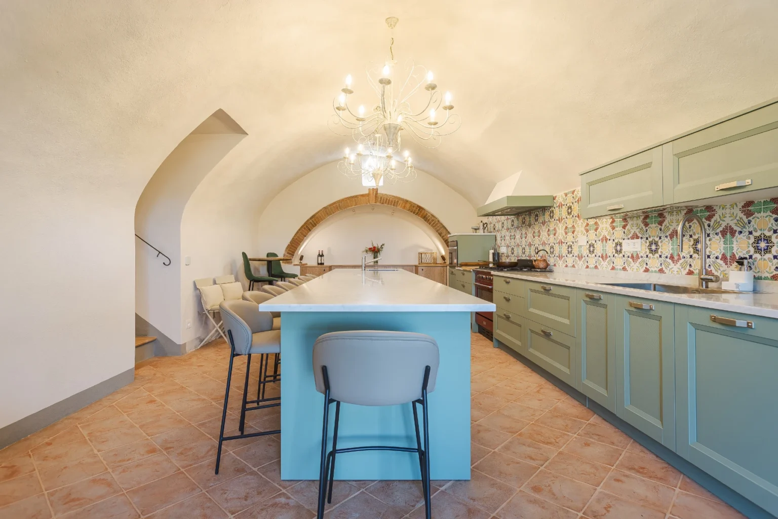 Modern kitchen interior featuring a large island with a light blue base, beige bar stools, and a decorative chandelier. The kitchen showcases green cabinetry, a colorful tiled backsplash, and an arched ceiling design, creating a spacious and stylish cooking area. | Only on SopranoVillas