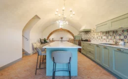 Modern kitchen interior featuring a large island with a light blue base, beige bar stools, and a decorative chandelier. The kitchen showcases green cabinetry, a colorful tiled backsplash, and an arched ceiling design, creating a spacious and stylish cooking area.