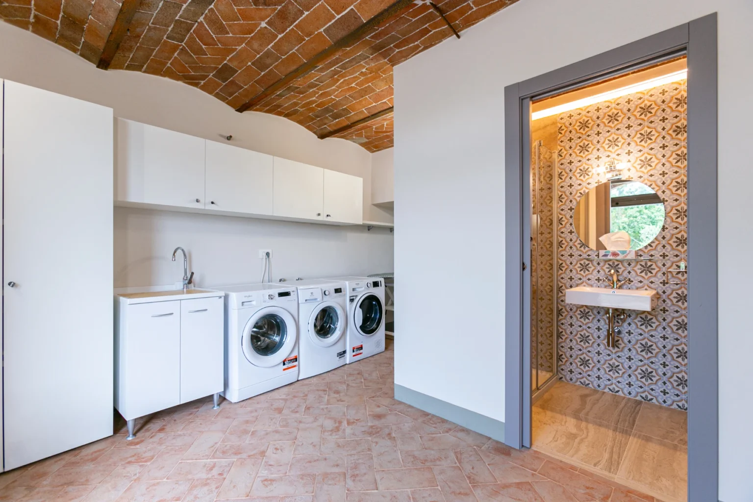 Modern laundry room featuring white appliances, a sleek sink, and a stylish bathroom entrance with patterned tile. The space has a rustic brick ceiling and ample storage, highlighting a functional and aesthetically pleasing design. | Only on SopranoVillas