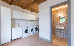 Modern laundry room featuring white appliances, a sleek sink, and a stylish bathroom entrance with patterned tile. The space has a rustic brick ceiling and ample storage, highlighting a functional and aesthetically pleasing design.