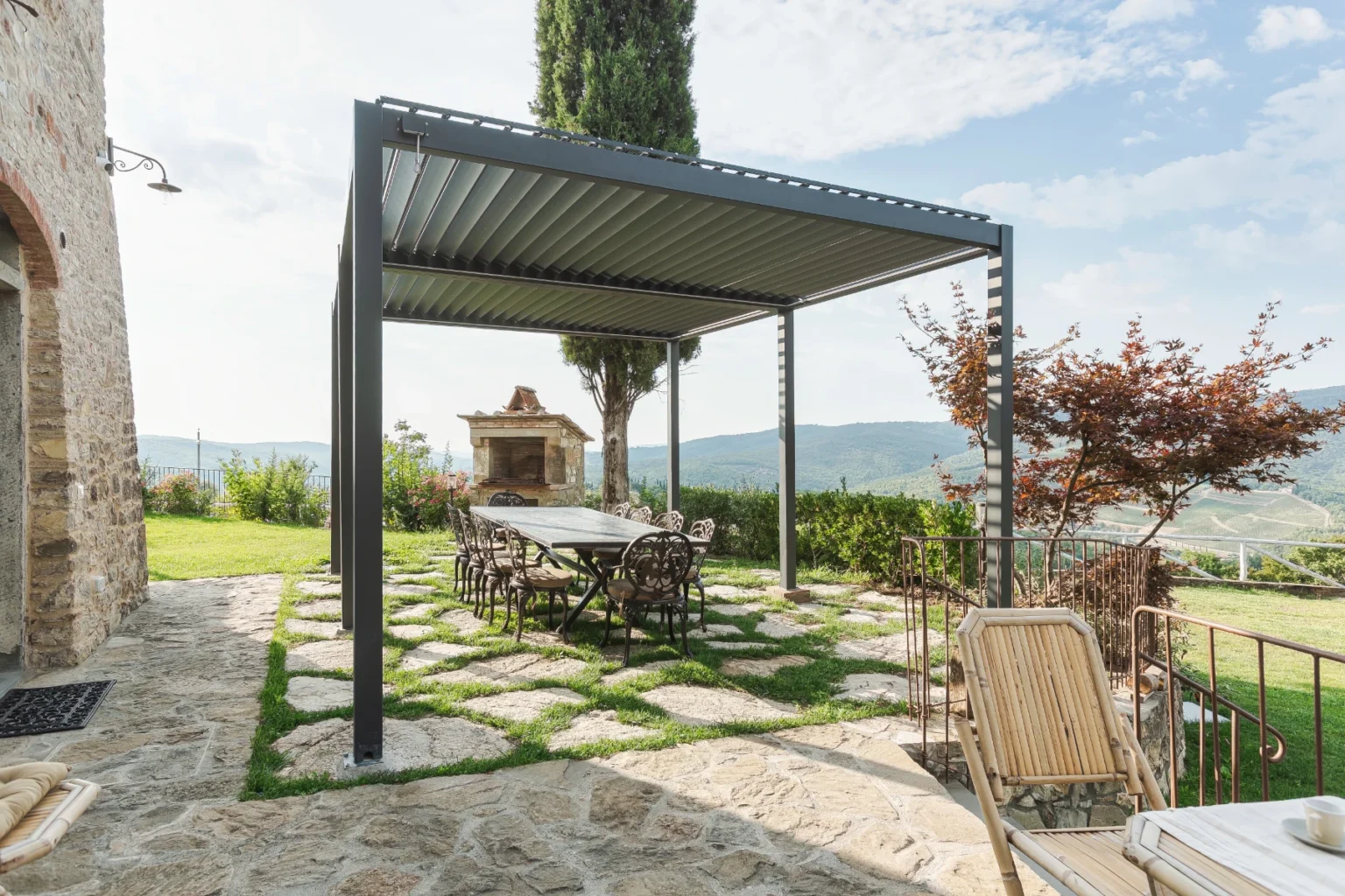 Outdoor dining area with a modern pergola, stone pathway, and a scenic view of hills and greenery. The setup includes a long table with wrought iron chairs and a stone barbecue grill nearby, ideal for gatherings and alfresco dining. | Only on SopranoVillas