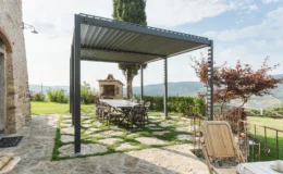Outdoor dining area with a modern pergola, stone pathway, and a scenic view of hills and greenery. The setup includes a long table with wrought iron chairs and a stone barbecue grill nearby, ideal for gatherings and alfresco dining.