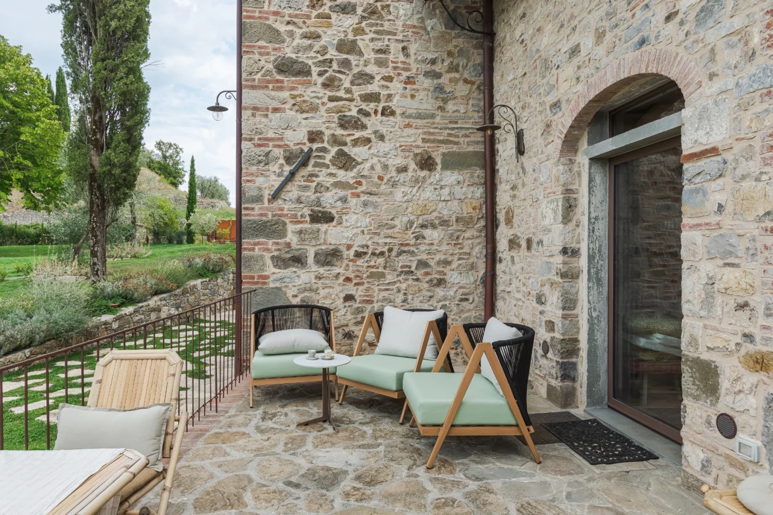 Outdoor seating area featuring modern chairs and a small table, set against a rustic stone wall with lush greenery in the background. Ideal for relaxation in a tranquil garden setting. | Only on SopranoVillas