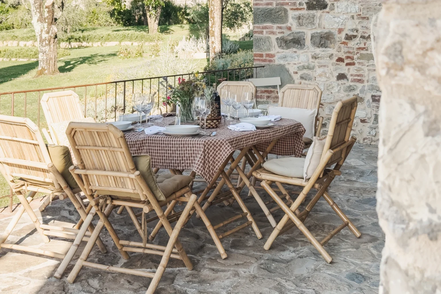Outdoor dining setup featuring a wooden table adorned with a brown checkered tablecloth, surrounded by stylish bamboo chairs with cushions. The table is elegantly set with plates, wine glasses, and a floral centerpiece, all situated on a stone patio with a lush green landscape in the background. | Only on SopranoVillas