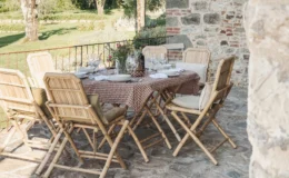 Outdoor dining setup featuring a wooden table adorned with a brown checkered tablecloth, surrounded by stylish bamboo chairs with cushions. The table is elegantly set with plates, wine glasses, and a floral centerpiece, all situated on a stone patio with a lush green landscape in the background.