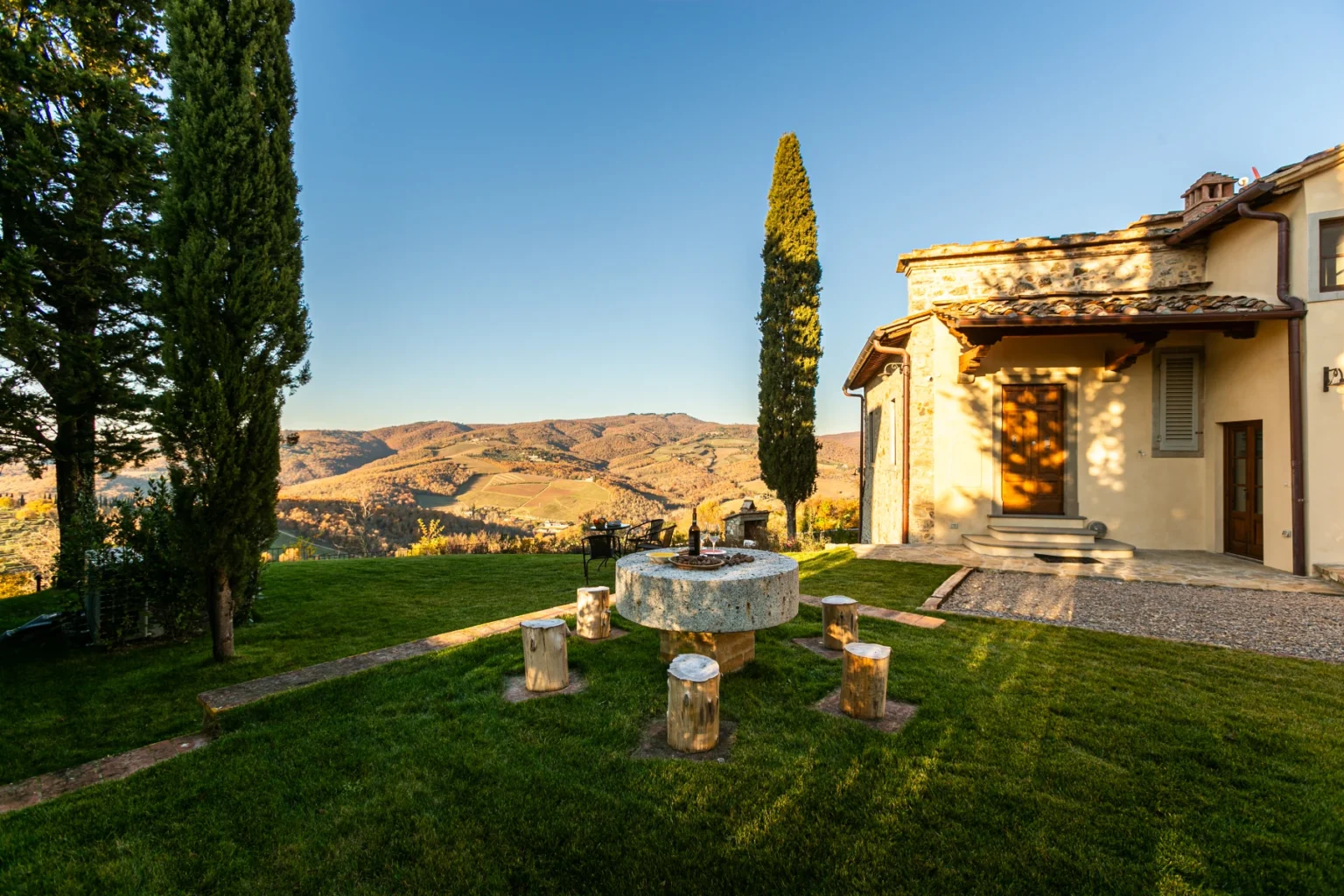 Scenic view of a rustic villa in Tuscany, featuring lush green grass, a stone fire pit surrounded by wooden stools, and rolling hills in the background under a clear blue sky. | Only on SopranoVillas