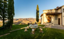 Scenic view of a rustic villa in Tuscany, featuring lush green grass, a stone fire pit surrounded by wooden stools, and rolling hills in the background under a clear blue sky.