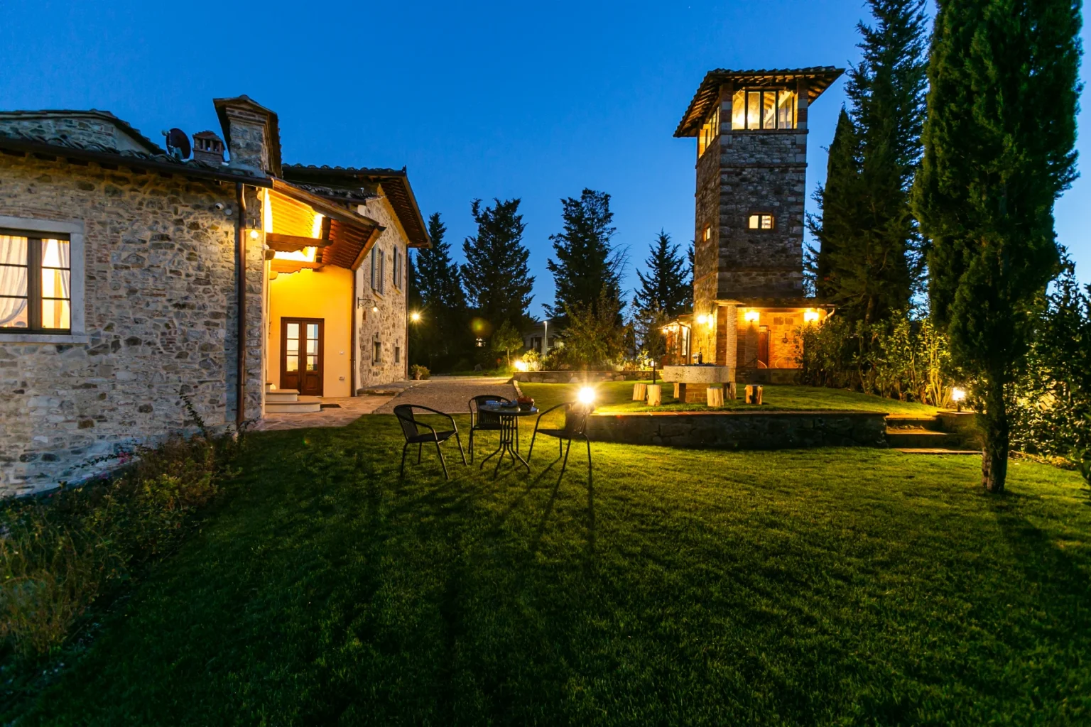 Stone villa with a tower at dusk, featuring a well-maintained lawn, outdoor seating, and illuminated pathways amidst lush greenery. Ideal for a tranquil getaway in a picturesque setting. | Only on SopranoVillas