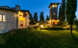 Stone villa with a tower at dusk, featuring a well-maintained lawn, outdoor seating, and illuminated pathways amidst lush greenery. Ideal for a tranquil getaway in a picturesque setting.