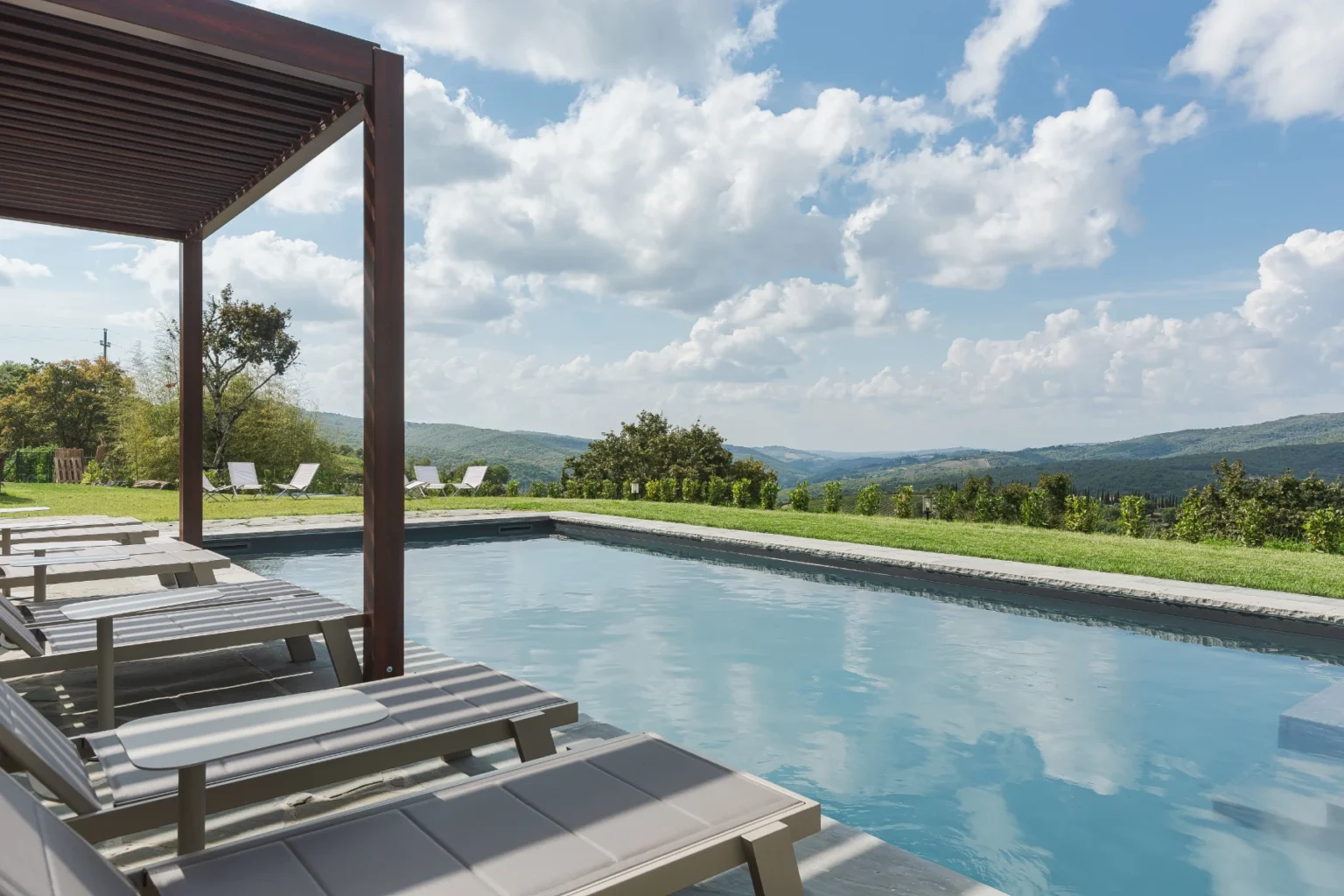 Serene outdoor swimming pool with lounge chairs, surrounded by lush green hills and a bright blue sky with fluffy clouds. Ideal for relaxation and enjoying nature. | Only on SopranoVillas