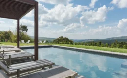 Serene outdoor swimming pool with lounge chairs, surrounded by lush green hills and a bright blue sky with fluffy clouds. Ideal for relaxation and enjoying nature.