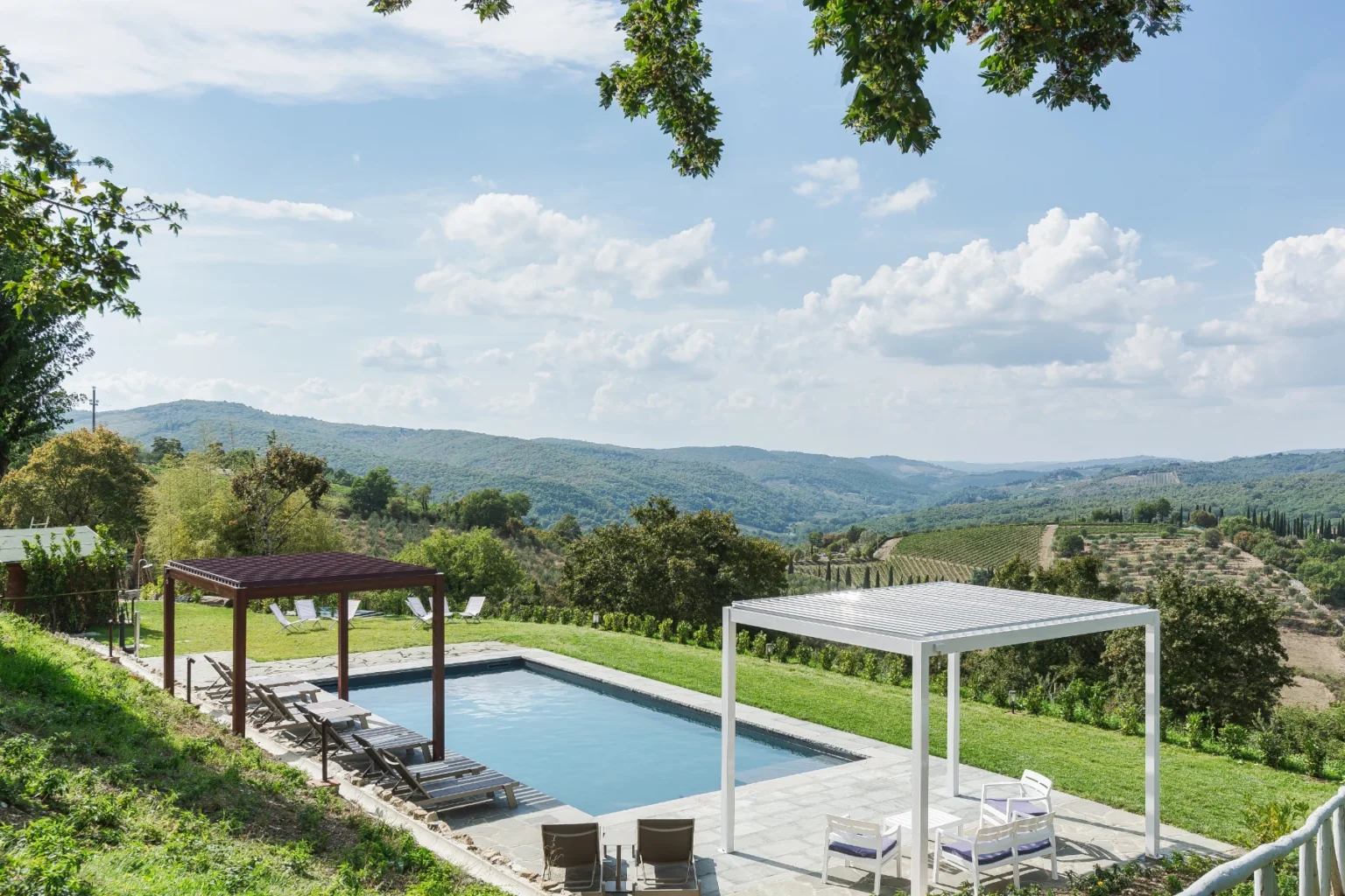 Scenic view of a modern swimming pool surrounded by lush greenery and rolling hills, featuring shaded lounge areas and a clear blue sky with scattered clouds, ideal for relaxation and outdoor enjoyment. | Only on SopranoVillas