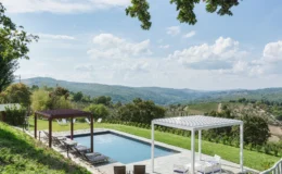 Scenic view of a modern swimming pool surrounded by lush greenery and rolling hills, featuring shaded lounge areas and a clear blue sky with scattered clouds, ideal for relaxation and outdoor enjoyment.
