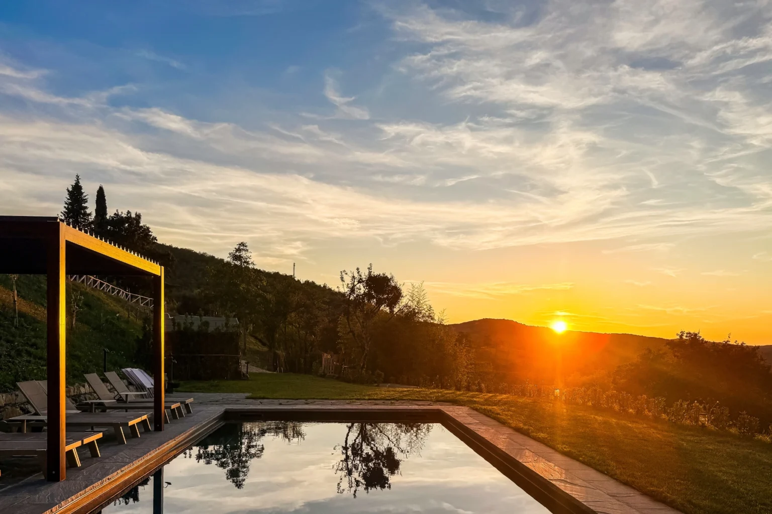 Scenic sunset view over a tranquil pool surrounded by lush greenery and hills, featuring a wooden pergola and lounge chairs for relaxation. | Only on SopranoVillas
