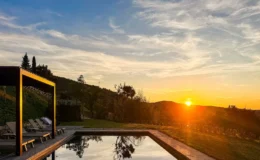 Scenic sunset view over a tranquil pool surrounded by lush greenery and hills, featuring a wooden pergola and lounge chairs for relaxation.