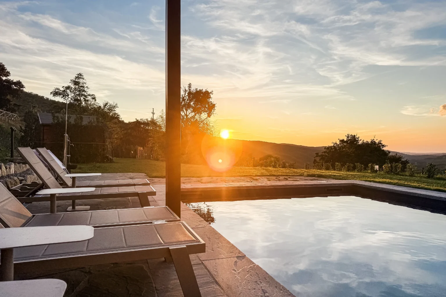 Serene sunset view over a swimming pool with lounge chairs, surrounded by lush greenery and rolling hills, showcasing a tranquil outdoor relaxation space. | Only on SopranoVillas