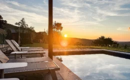 Serene sunset view over a swimming pool with lounge chairs, surrounded by lush greenery and rolling hills, showcasing a tranquil outdoor relaxation space.