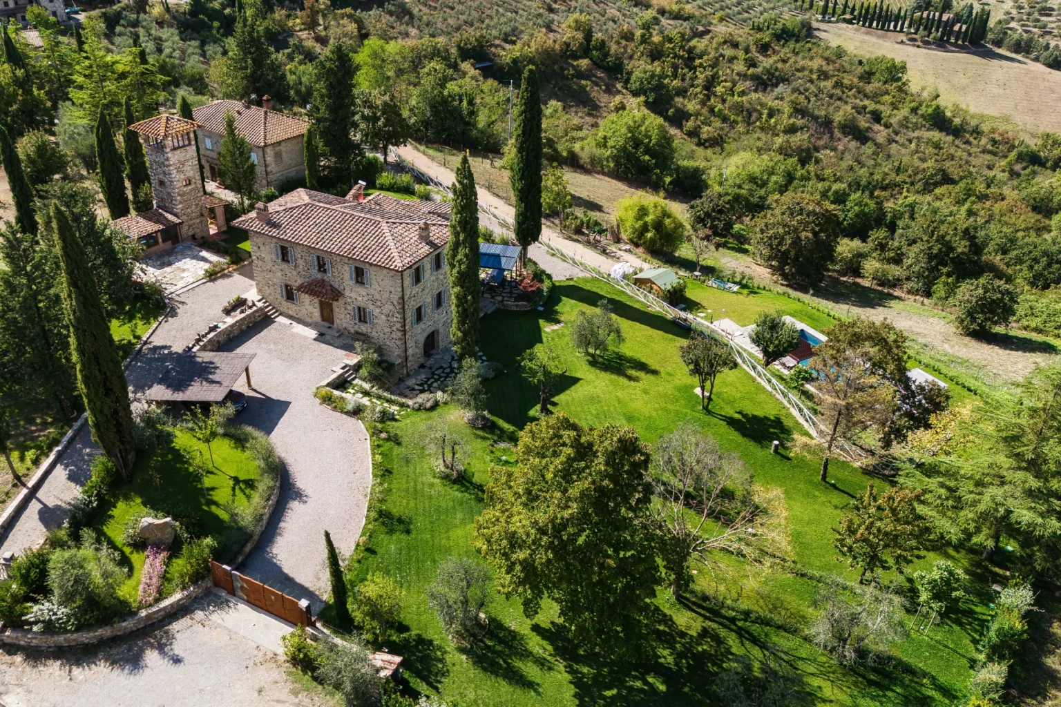 Aerial view of a charming stone villa surrounded by lush greenery and landscaped gardens, featuring a spacious driveway, tall cypress trees, and a swimming pool area in the background, set in a picturesque countryside landscape. | Only on SopranoVillas