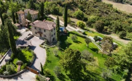Aerial view of a charming stone villa surrounded by lush greenery and landscaped gardens, featuring a spacious driveway, tall cypress trees, and a swimming pool area in the background, set in a picturesque countryside landscape.
