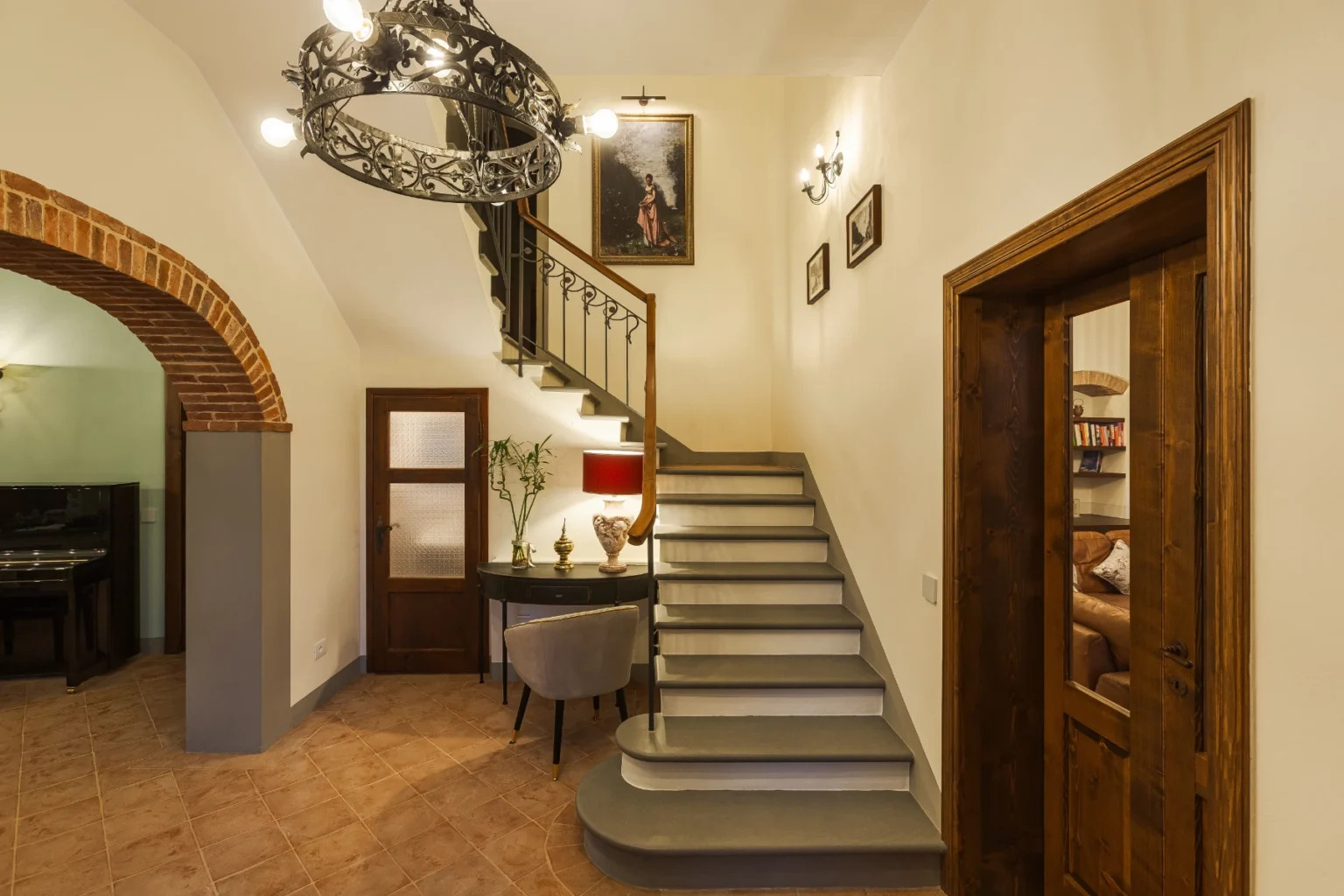 Elegant entryway featuring a decorative staircase, a vintage chandelier, and stylish decor. The space includes a console table with a lamp and a potted plant, complemented by warm lighting and artistic wall decor, creating a welcoming atmosphere. | Only on SopranoVillas