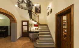 Elegant entryway featuring a decorative staircase, a vintage chandelier, and stylish decor. The space includes a console table with a lamp and a potted plant, complemented by warm lighting and artistic wall decor, creating a welcoming atmosphere.