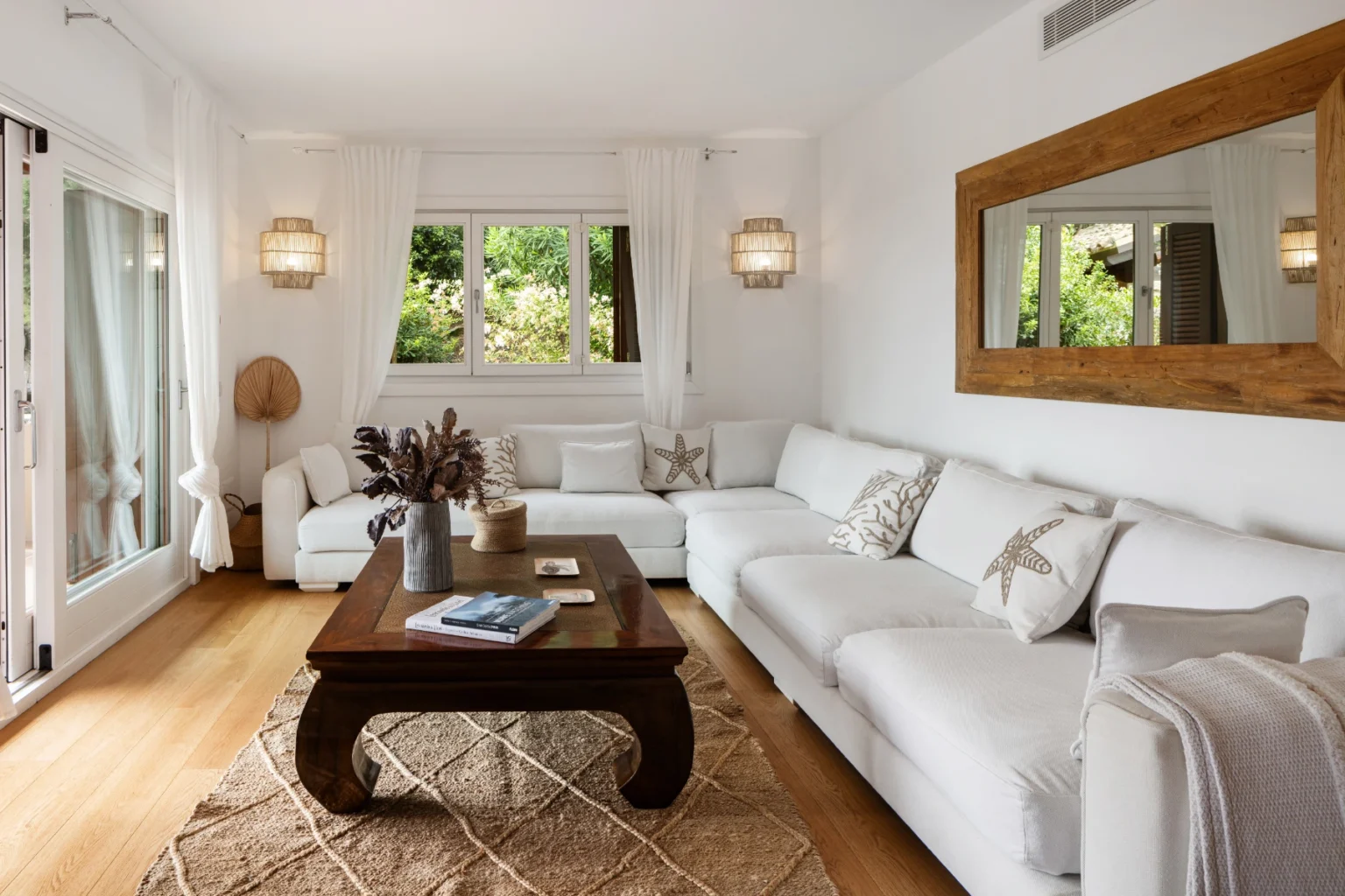 Bright and airy living room featuring a white sectional sofa, wooden coffee table, and natural light from large windows, complemented by decorative plants and a stylish mirror. | Only on SopranoVillas