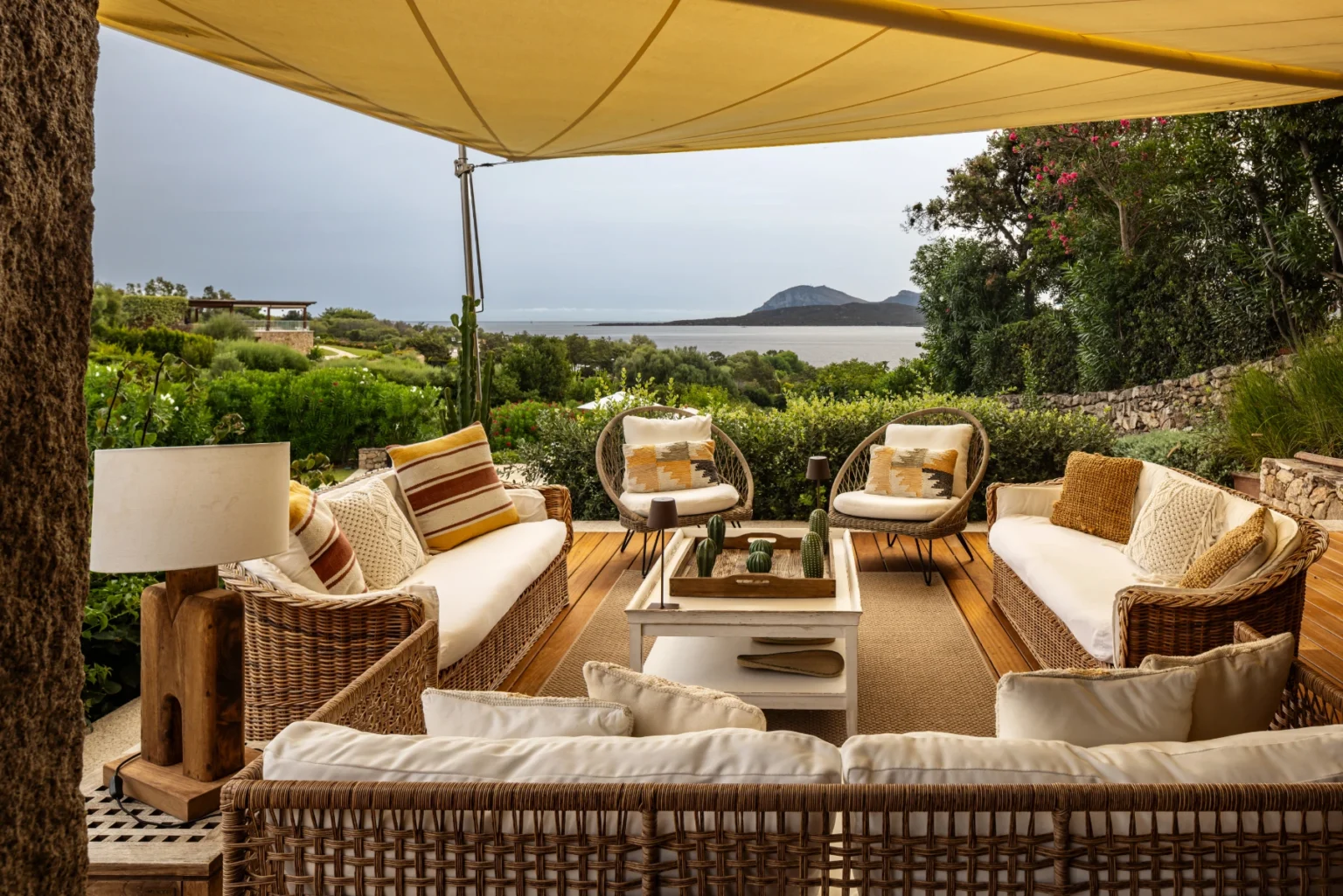Cozy outdoor seating area with comfortable wicker furniture, plush cushions, and a coffee table, set under a large canopy, overlooking a scenic view of lush greenery and a distant coastline. | Only on SopranoVillas