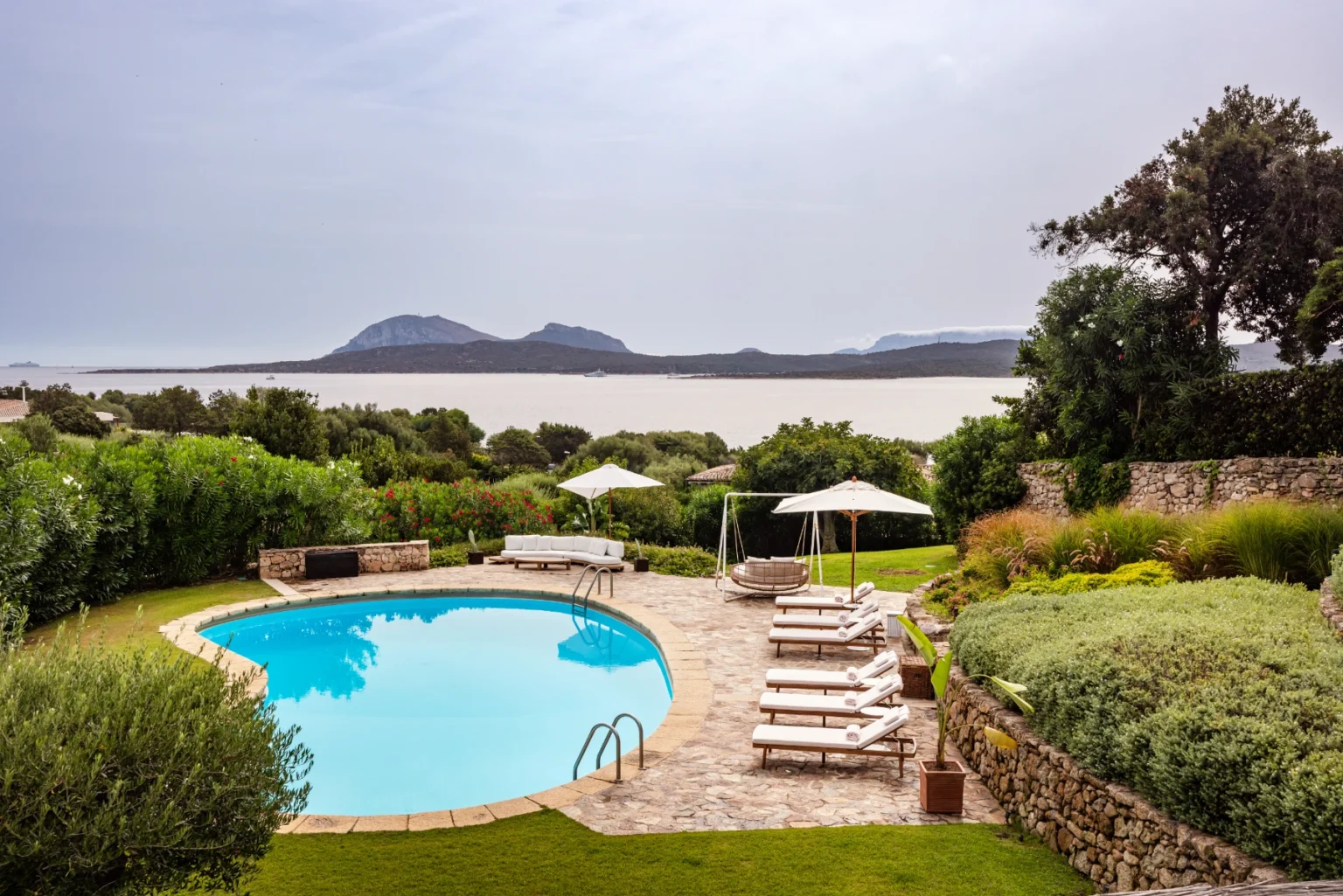 Scenic outdoor pool surrounded by lush greenery and lounge chairs, with a view of mountains and a calm lake in the background. Ideal for relaxation and leisure in a tranquil setting. | Only on SopranoVillas
