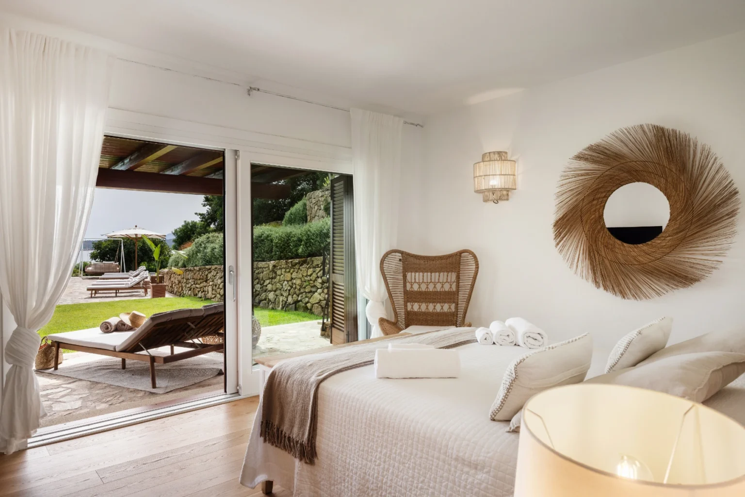 Bright and airy bedroom featuring a king-sized bed, elegant decor, and large glass doors leading to a lush outdoor patio with lounging furniture. Natural light floods the space, enhancing the serene atmosphere. | Only on SopranoVillas
