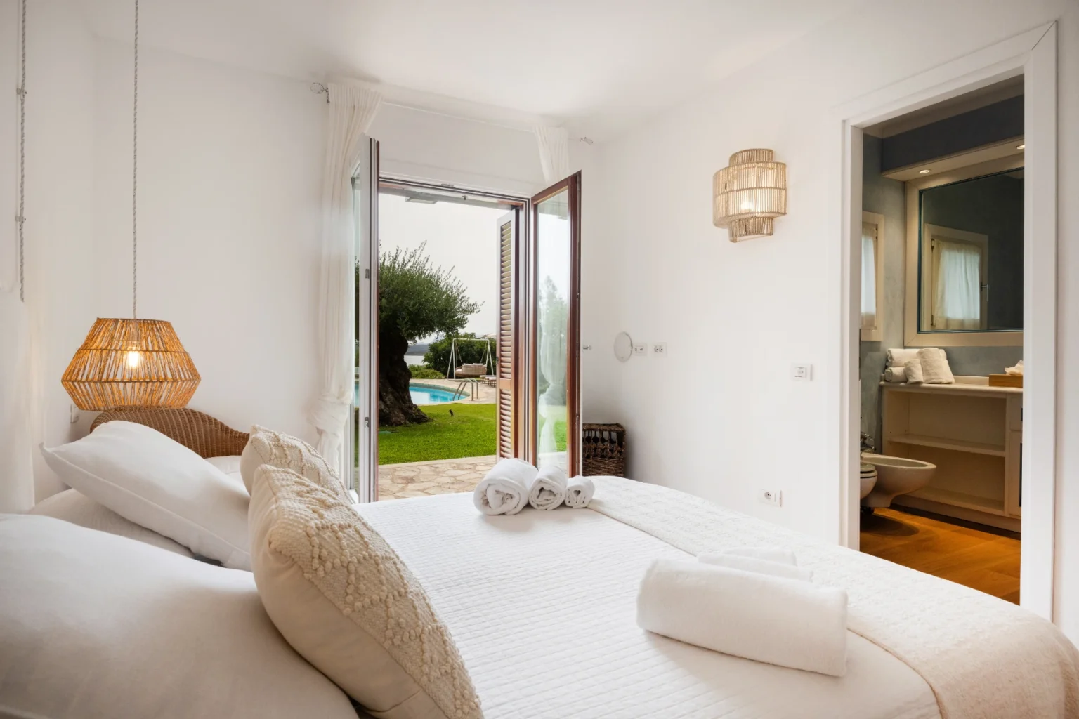 Bright and airy bedroom with a double bed, decorative pillows, and natural light streaming through open doors leading to a garden and pool area. Features a cozy atmosphere with stylish lighting and an adjoining bathroom visible in the background. Ideal for relaxation and comfort. | Only on SopranoVillas