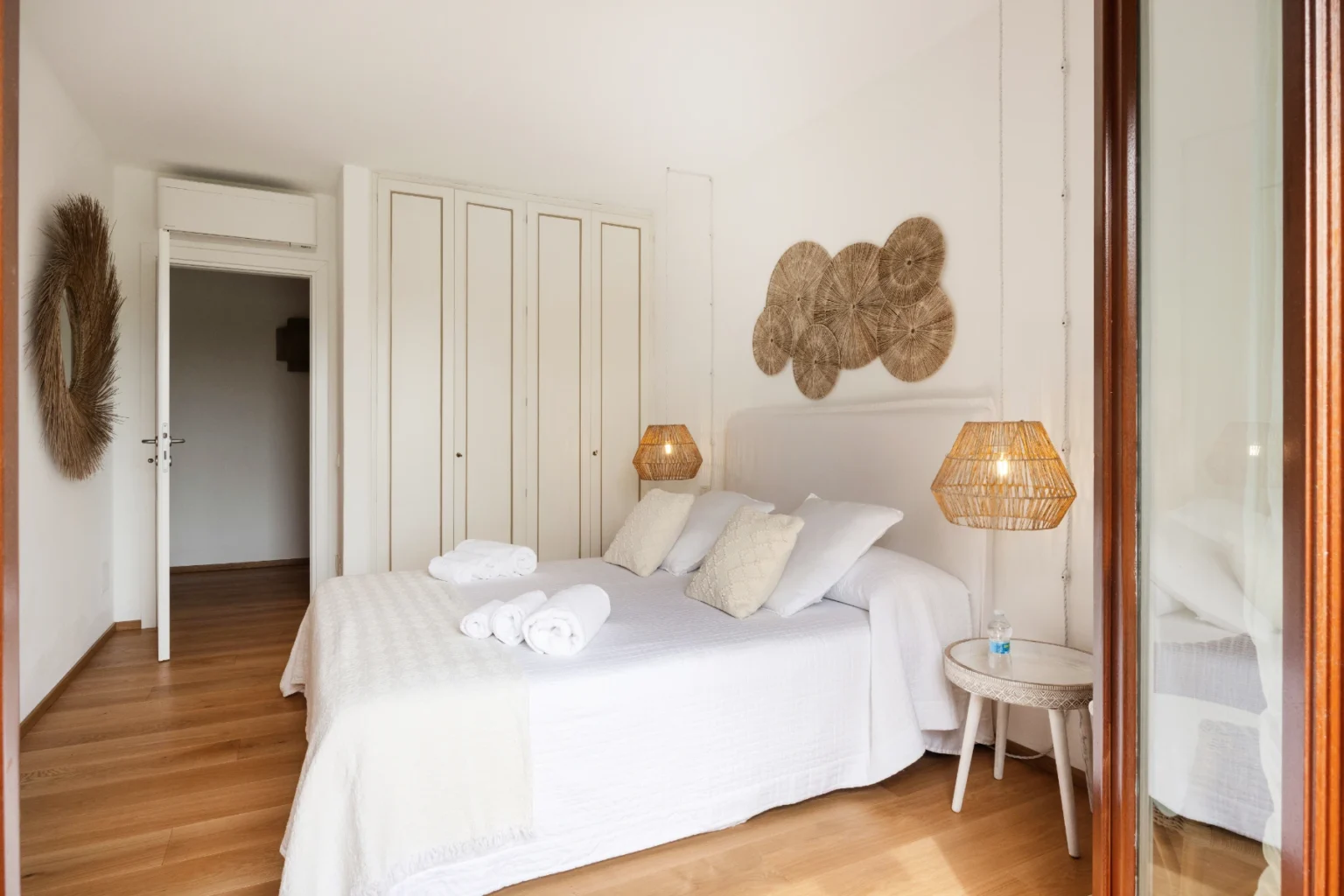 Cozy bedroom interior featuring a double bed with white linens, wooden flooring, and stylish wicker lamps. The room includes a modern closet and decorative wall art, creating a serene and inviting atmosphere. | Only on SopranoVillas