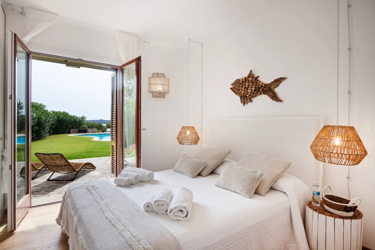 Bright and airy bedroom with white decor, featuring a cozy bed with decorative pillows and towels, wicker light fixtures, and a fish wall art. The room opens to a lush outdoor view with a swimming pool and lounge chairs, creating a serene atmosphere. | Only on SopranoVillas