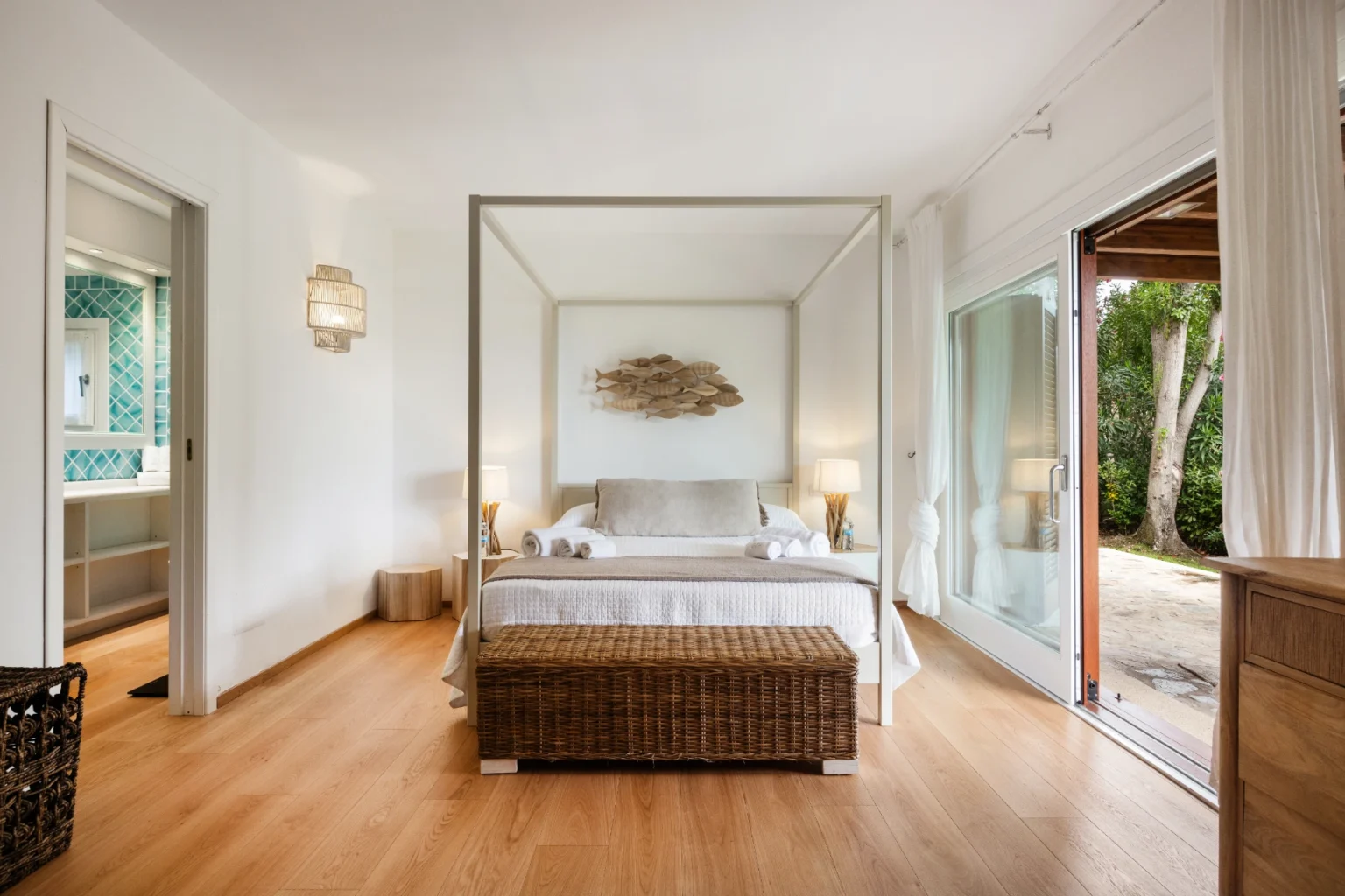 Modern bedroom with a four-poster bed, wooden flooring, and natural light streaming in through large sliding glass doors. The room features minimalist decor, a woven bench at the foot of the bed, and a serene atmosphere, perfect for relaxation. | Only on SopranoVillas