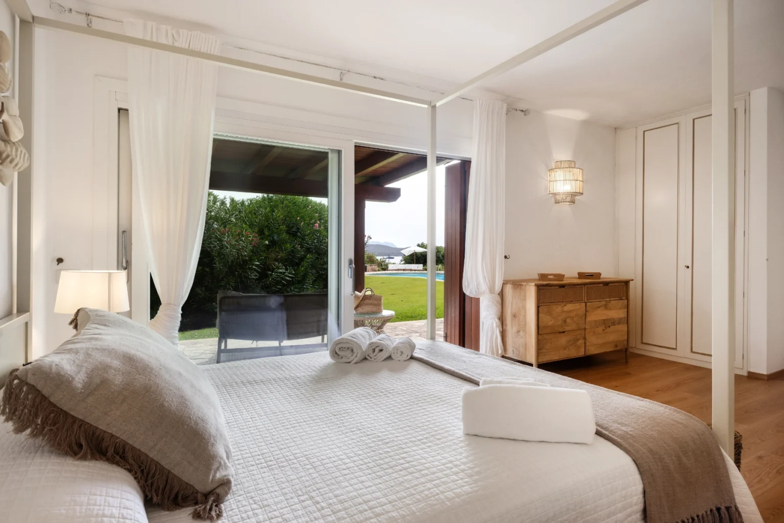 Bright and airy bedroom featuring a four-poster bed, elegant decor, and large sliding glass doors leading to a garden view. The room includes a wooden dresser, bedside lamp, and soft textiles for a cozy atmosphere. Ideal for relaxation and comfort. | Only on SopranoVillas