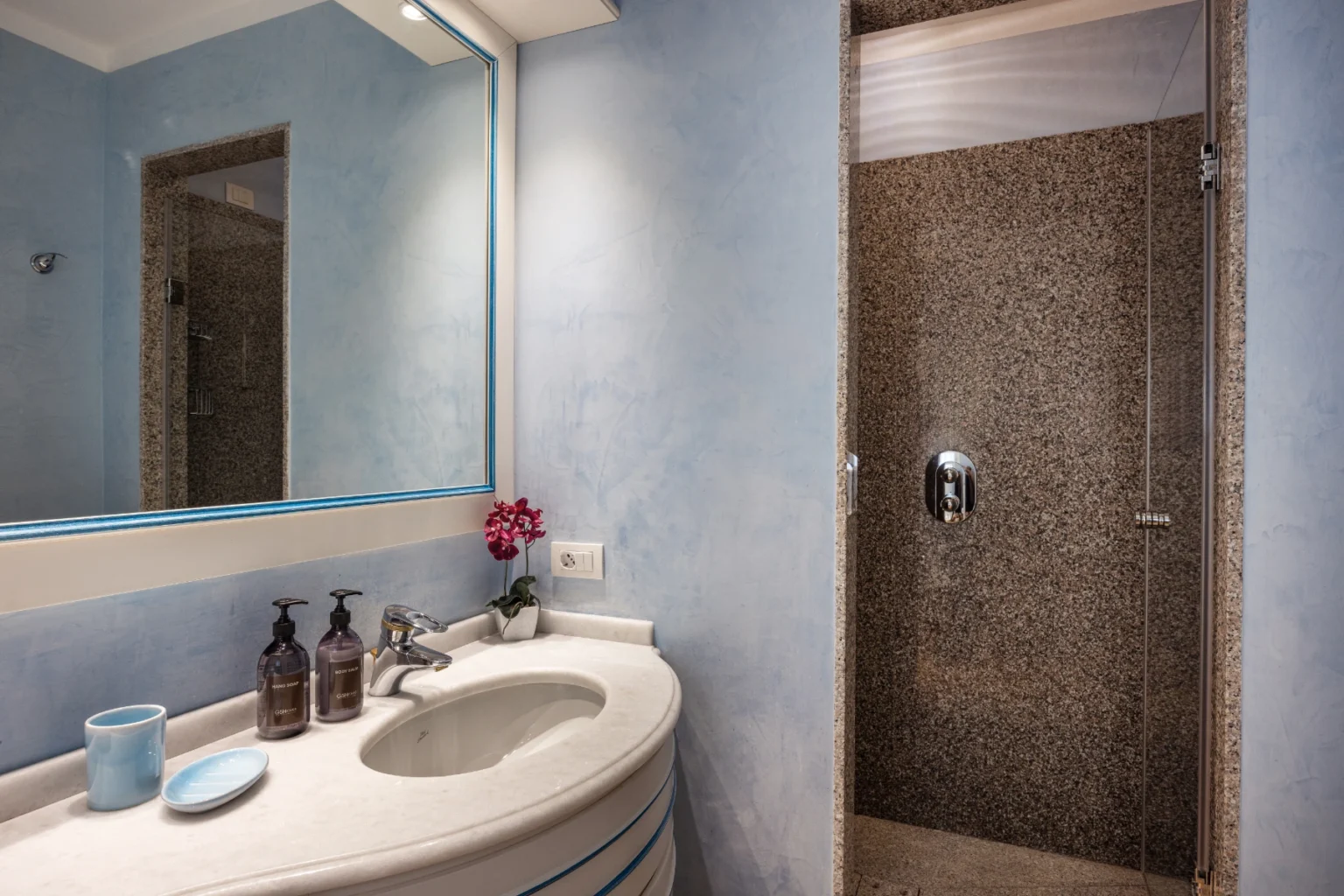 Modern bathroom featuring a blue wall design, a large mirror, a curved sink with toiletries, and a stylish shower with mosaic tile. | Only on SopranoVillas