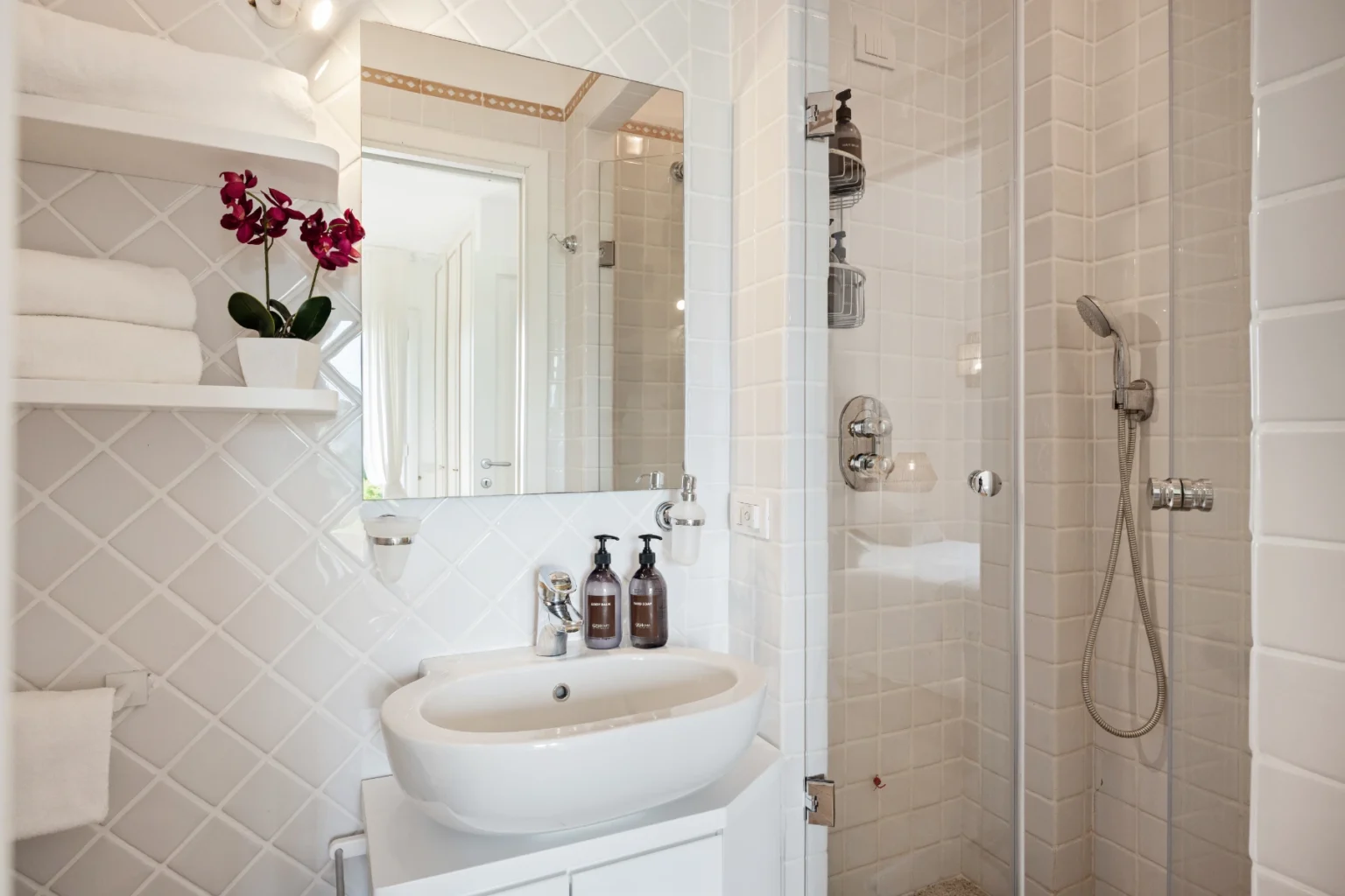 Modern bathroom with a sleek design featuring a shower, a stylish sink, white tiles, and decorative elements like a potted orchid and neatly arranged towels on shelves. Ideal for showcasing contemporary bathroom aesthetics. | Only on SopranoVillas