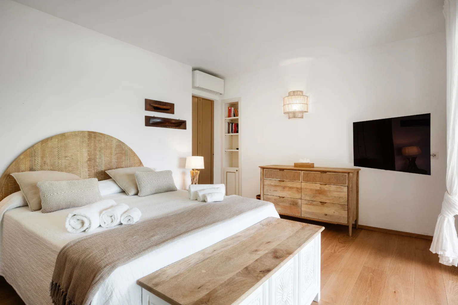 Modern bedroom featuring a wooden headboard, neatly made bed with decorative pillows, a stylish dresser, and a flat-screen TV on the wall, all set against a bright, minimalist decor. | Only on SopranoVillas