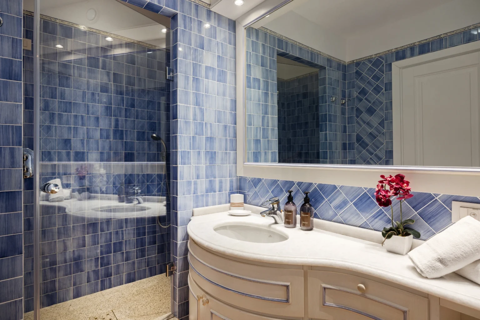 Modern bathroom featuring blue tiled walls, a glass shower enclosure, a stylish vanity with a white countertop, and decorative elements like bottles and a flower arrangement. | Only on SopranoVillas