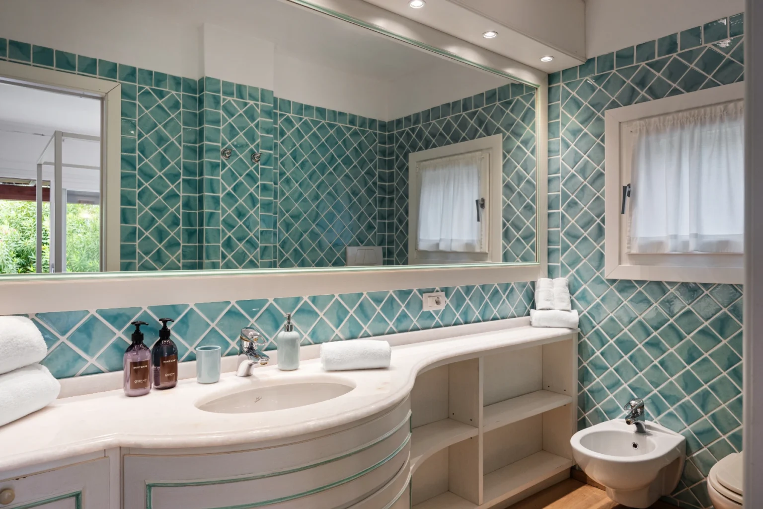 Modern bathroom featuring turquoise diamond-patterned tiles, a sleek countertop with a sink, a bidet, and a large mirror. Natural light enters through a window, enhancing the spacious feel of the room. | Only on SopranoVillas