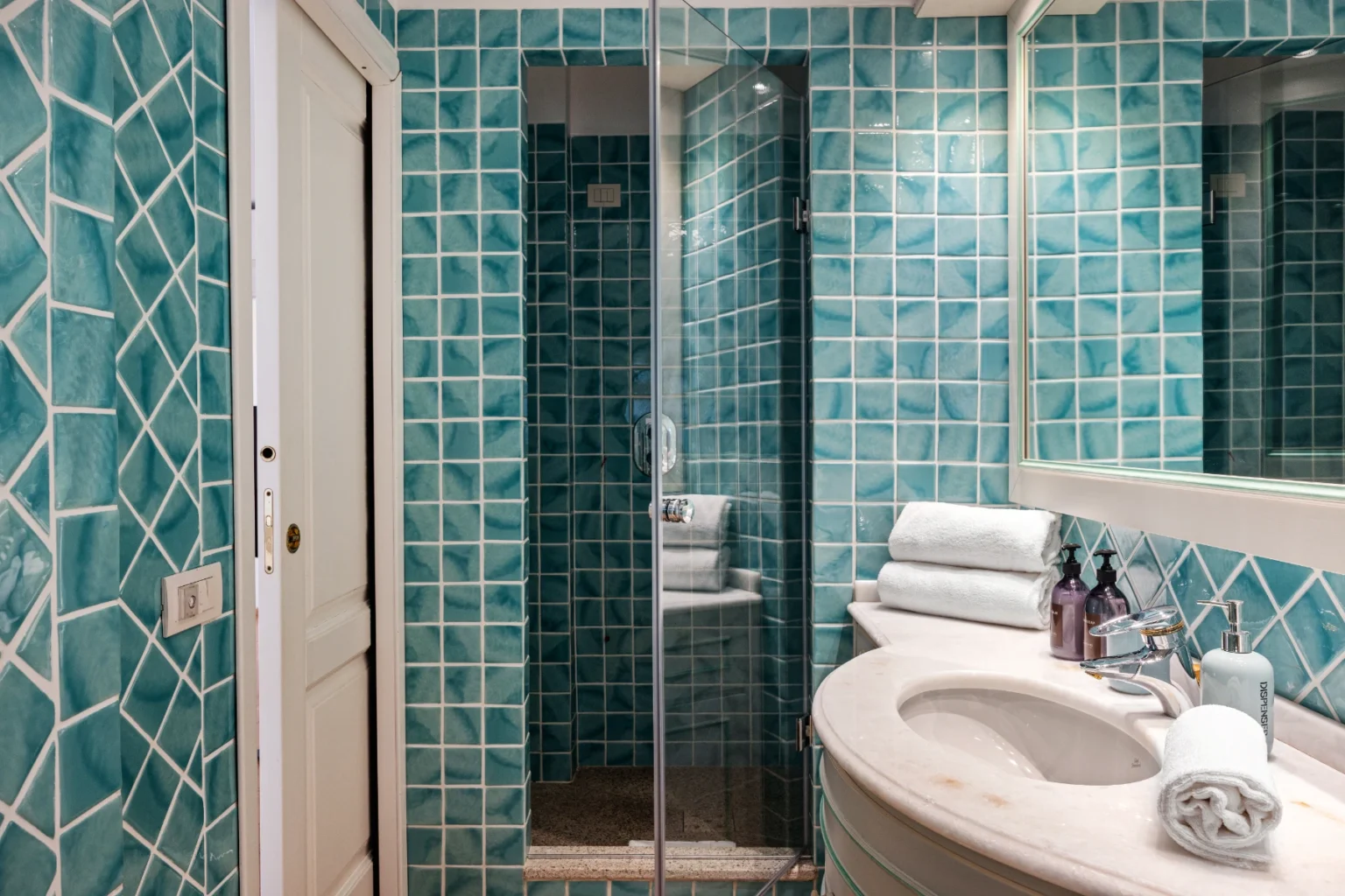 Modern bathroom featuring turquoise geometric tile walls, a glass shower enclosure, a white sink with neatly arranged towels, and contemporary fixtures. | Only on SopranoVillas
