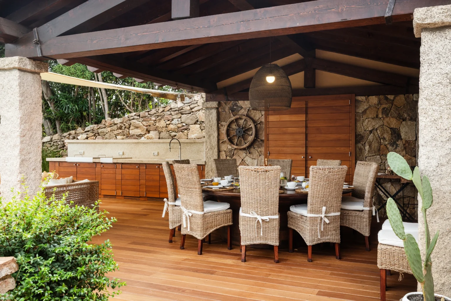 Outdoor dining area featuring a wooden table set with chairs, surrounded by stone walls and lush greenery. The space is shaded by a roof, creating a cozy atmosphere for gatherings. | Only on SopranoVillas