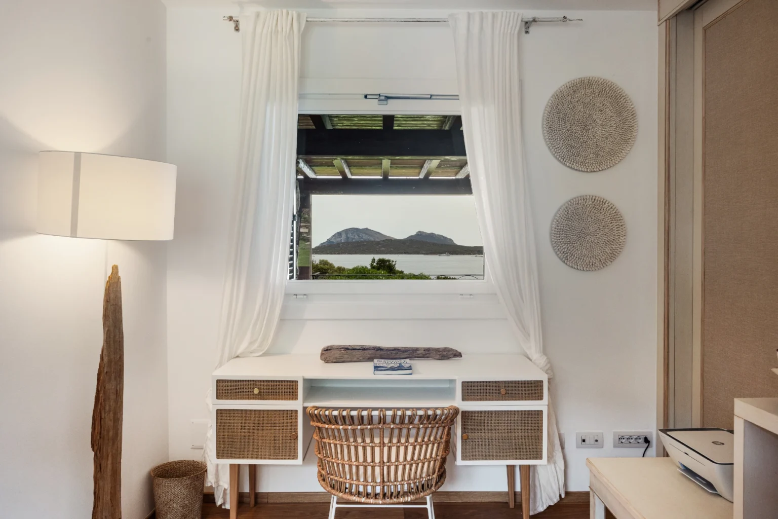 Modern home office setup featuring a white desk and a wicker chair, adorned with decorative wall hangings and a large window showcasing a scenic view of mountains and water. Natural light illuminates the space, enhancing its serene atmosphere. | Only on SopranoVillas
