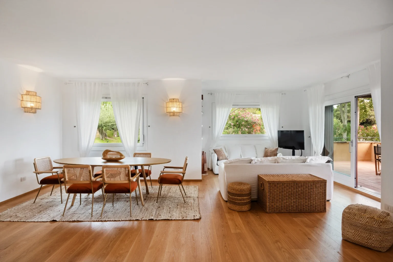 Bright and airy modern living room featuring a wooden dining table with eight chairs, stylish pendant lights, and large windows offering a view of greenery. The space includes a comfortable white sofa, a woven rug, and a television, creating a welcoming atmosphere. | Only on SopranoVillas