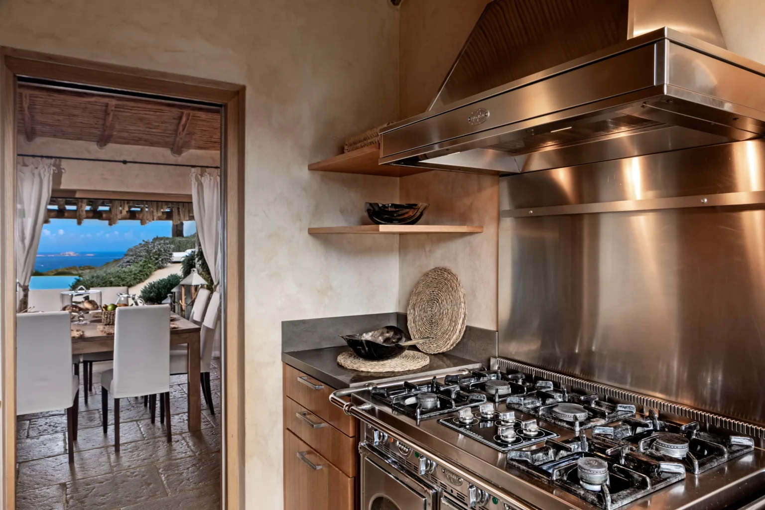 Luxury villa kitchen in Italy featuring high-end stainless steel appliances, wooden cabinetry, and a scenic view of the dining area and landscape beyond. Ideal for gourmet cooking and entertaining. | Only on SopranoVillas