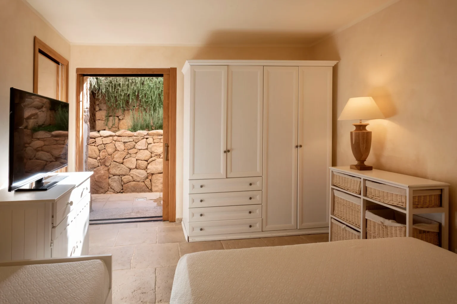 Luxury villa bedroom in Italy featuring a serene design with a white wardrobe, dresser, and a view of the outdoor stone wall, ideal for vacation rentals. | Only on SopranoVillas