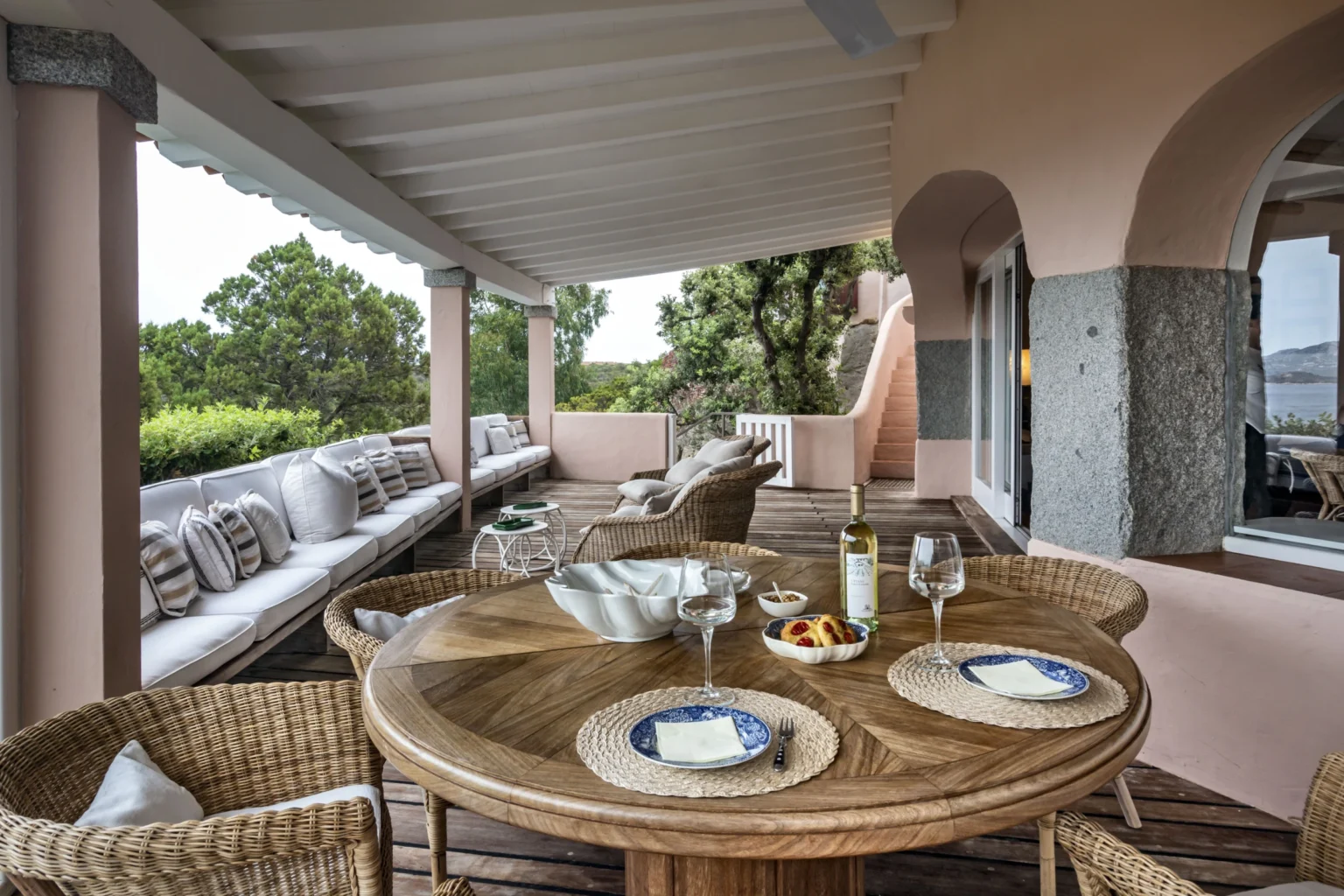 Elegant outdoor dining area featuring a round wooden table set with plates and glasses, surrounded by comfortable wicker chairs. A cozy lounge area with plush cushions is visible in the background, nestled among lush greenery. Ideal for alfresco dining and relaxation. | Only on SopranoVillas